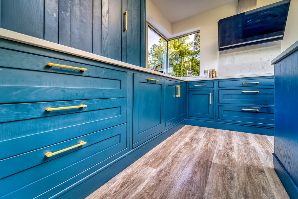 Run of blue kitchen drawers with brass handles beneath a corner window